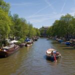 Amsterdam Sehenswürdigkeiten: Museen, Tickets und Grachten Gracht in der Amsterdamer Innenstadt mit Booten, Giebelhäusern und Baumreihen im Sommer