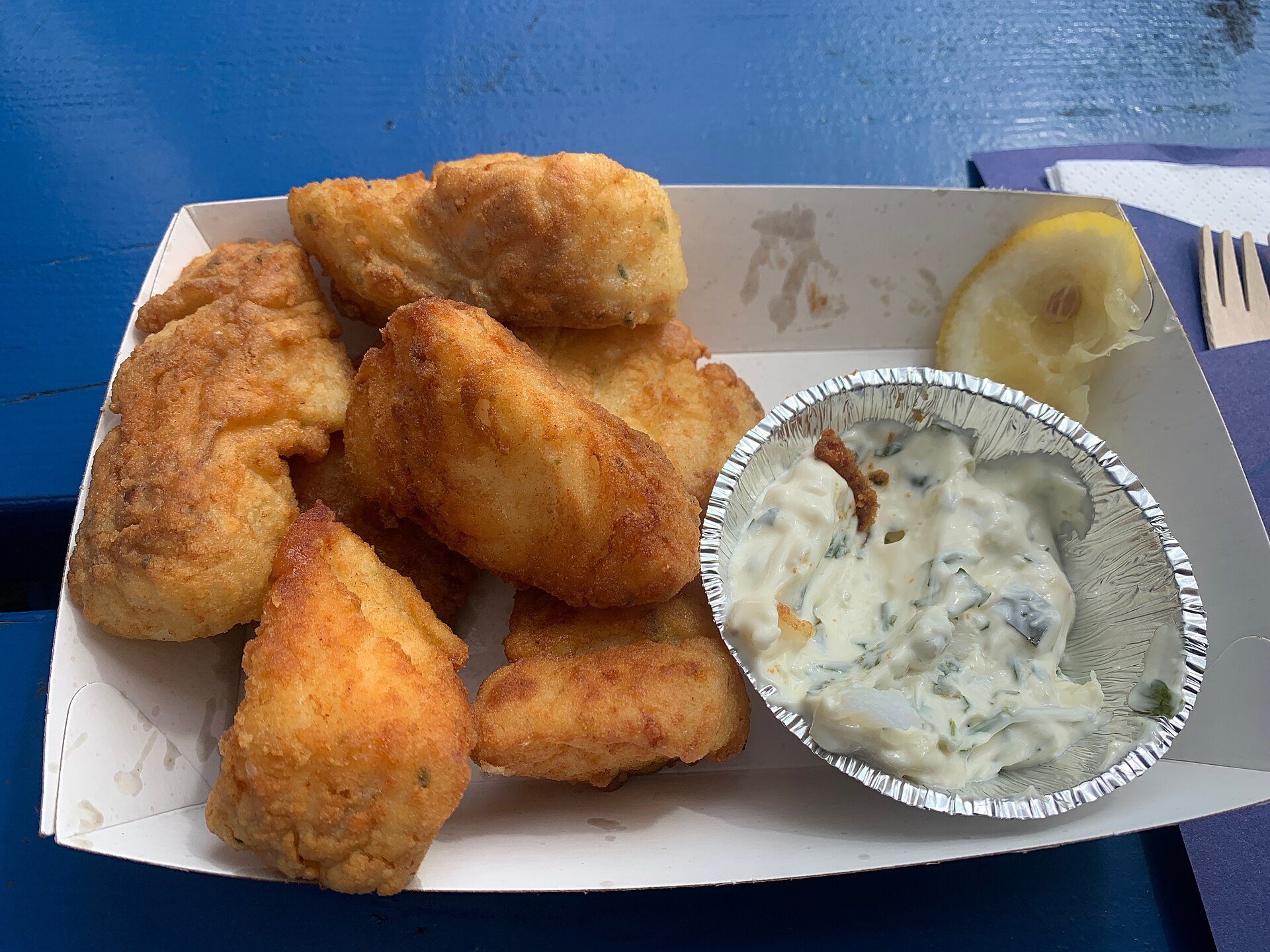 Portion Kibbeling mit Kräutersauce und Zitronenspalte in der Pappschale
